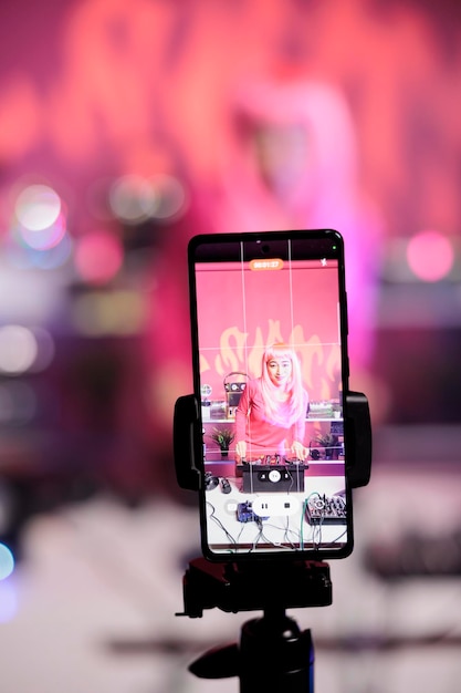 Artiste joyeux aux cheveux roses debout à la table de dj mélangeant une chanson avec des platines devant la caméra tout en enregistrant une vidéo avec un téléphone. Femme souriante faisant des performances dans une discothèque avec équipement audio