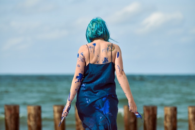 Artiste de performance artistique femme aux cheveux bleus en robe bleu foncé enduite de peinture à la gouache bleue et danse sur la plage, vue arrière. Peinture corporelle, concept d'art corporel, art de la performance expressif en plein air