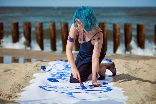 Artiste de performance artistique femme aux cheveux bleus en robe bleu foncé enduite de peinture à la gouache bleue avec de larges traits sur une grande toile sur la plage. Art de la performance expressif, art contemporain en plein air