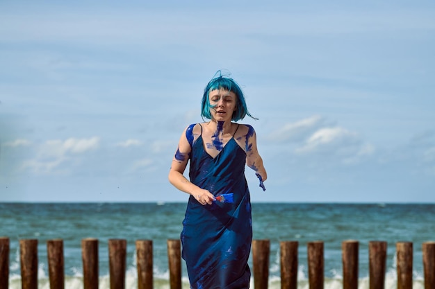 Artiste de performance artistique femme aux cheveux bleus en robe bleu foncé enduite de peinture à la gouache bleue peignant et dansant sur la plage. Concept de peinture corporelle et d'art corporel, art de la performance expressif à l'extérieur