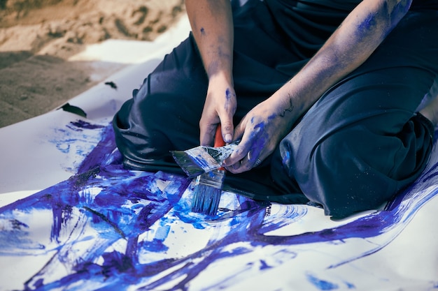 Artiste de performance féminine en robe bleu foncé enduite de peinture à la gouache bleue avec de larges traits sur une grande toile allongée sur la plage, gros plan. Performance expressive, art contemporain, plein air