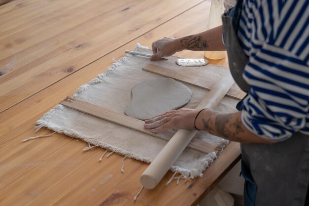 Une artiste potière en tablier utilise un rouleau à pâtisserie en bois pour dérouler de l'argile faisant des ustensiles faits à la main