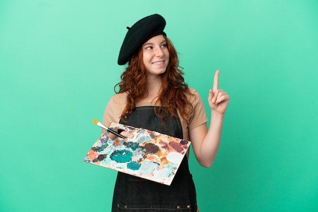 Artiste rousse adolescent tenant une palette isolée sur fond vert pointant vers une excellente idée