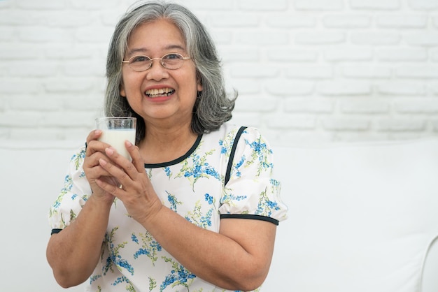 Asian Senior woman holding glass milk tout en se relaxant sur un canapé salon pour le bien-être à la retraite