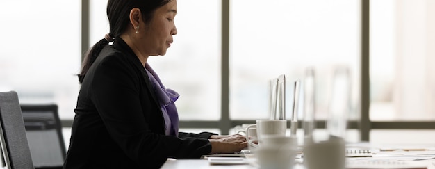 Asiatique d'âge moyen, heureuse, confiante, femme d'affaires prospère, secrétaire en costume formel, assise au bureau, tapant des informations sur un ordinateur portable seul dans une salle de bureau calme de l'entreprise.