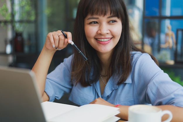 Asiatique belle femme en chemise bleue à l'aide d'ordinateur portable et de boire du café