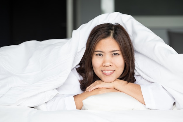 Asiatique femme allongée au bout du lit sous la couette et souriant