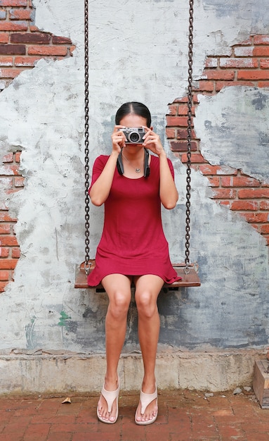 Asiatique Femme Prenant La Caméra Et Assis Sur La Balançoire Contre Le Vieux Fond De Mur De Brique