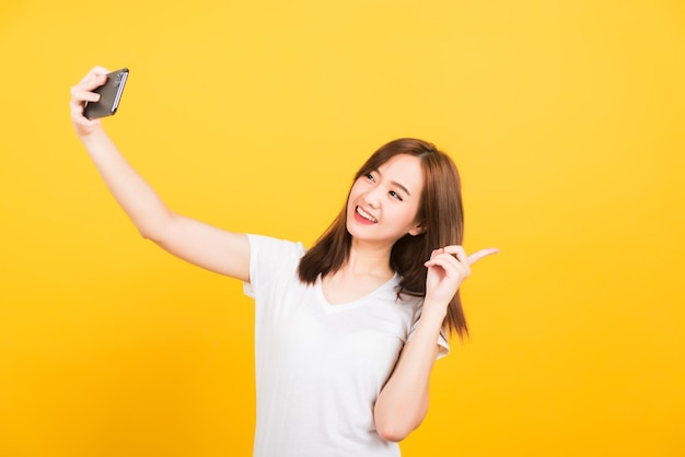 Asiatique heureux portrait belle jolie jeune femme adolescent souriant debout porter t-shirt mak selfie photo, appel vidéo sur smartphone, pointer le doigt vers l'arrière isolé, studio tourné fond jaune avec espace de copie