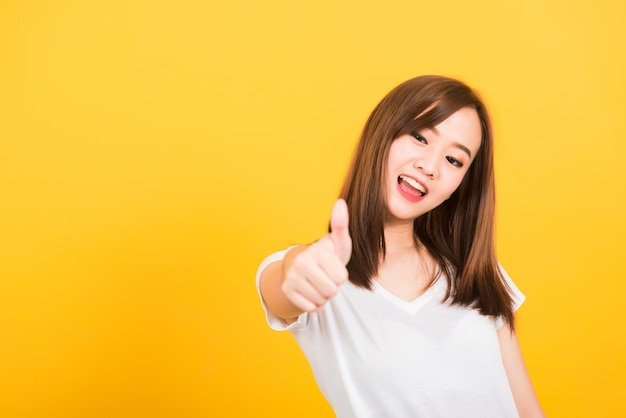 Asiatique heureux portrait belle jolie jeune femme adolescente debout porter un t-shirt montrant des gestes doigt pouce vers le haut regardant vers la caméra isolée, prise de vue en studio sur fond jaune avec espace de copie