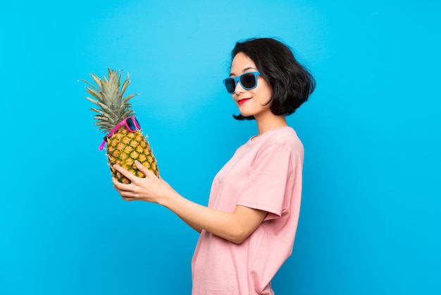 Asiatique jeune femme sur un mur bleu isolé, tenant un ananas avec des lunettes de soleil