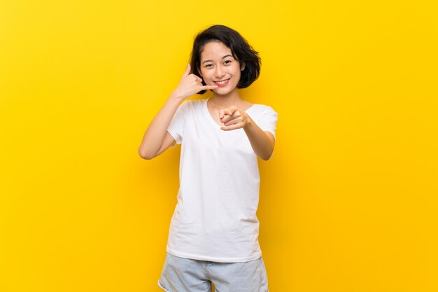 Asiatique jeune femme sur mur jaune isolé faisant un geste de téléphone et pointant le devant