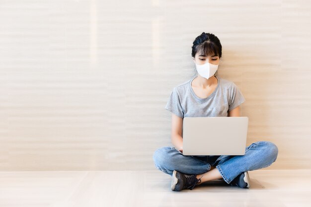 Asiatique jeune femme portant un masque de protection et assis sur le sol et travaillant à l'aide d'un ordinateur portable.