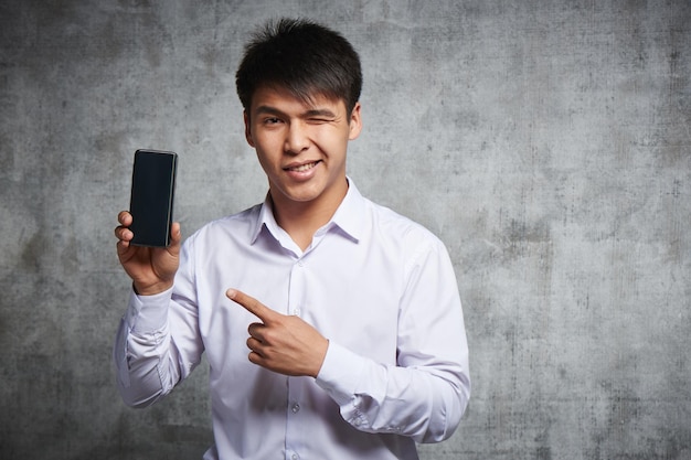 Asiatique joyeux pointant sur un téléphone portable avec un index faisant la publicité de quelque chose. Kazakh attrayant en chemise blanche isolé en studio