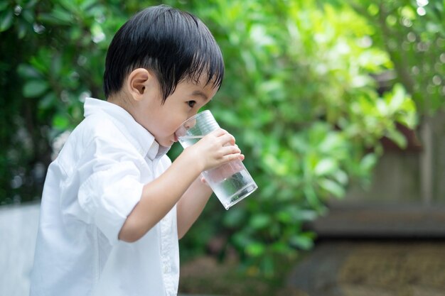 Asiatique mignon garçon porter chemise blanche, boire de l&#39;eau froide de verre dans le fond de la nature verte