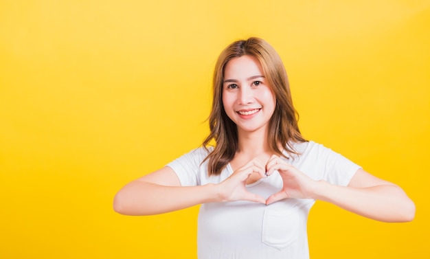 Asiatique thaïlandais heureux portrait belle jolie jeune femme sourire debout faire doigt coeur figure symbole forme signe avec deux mains et regardant la caméra, studio tourné fond jaune isolé avec espace de copie