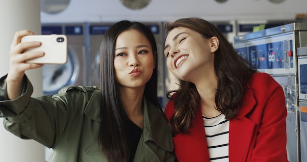 Asiatiques et caucasiennes jeunes belles filles souriant et posant pour l'appareil photo du smartphone tout en prenant une photo de selfie au service de blanchisserie. Jolies femmes faisant des photos de selfies avec téléphone dans les machines à laver.