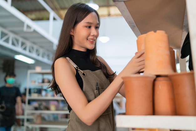 Asie femme céramiste avec tablier main arranger son produit de sculpture d'argile sur l'étagère en attente de cuisson dans une femme asiatique propriétaire d'un atelier de céramique de petite entreprise souriante fière de présenter sa nouvelle collection