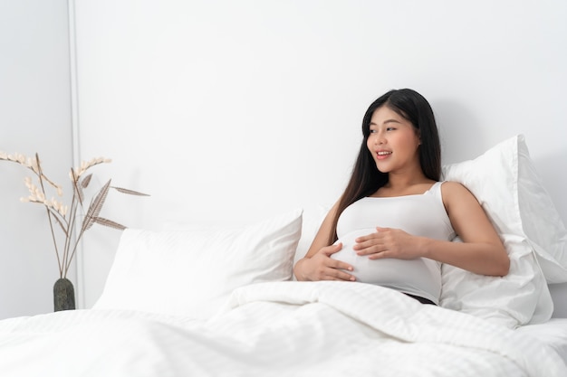 Asie femme enceinte heureuse est assise sur le lit et touche son ventre. Concept de grossesse, maternité, personnes et attente