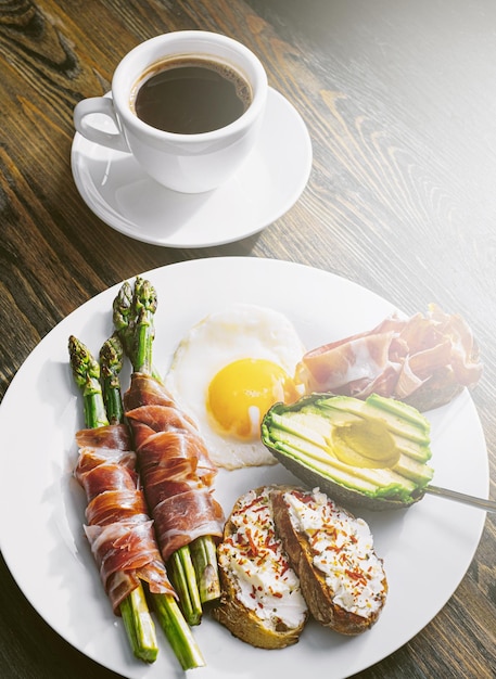 Asperges frites enveloppées dans une bruschetta d'œufs frits au jamon avec du fromage à pâte molle et de l'avocat sur une assiette blanche et une tasse de café fraîchement moulu