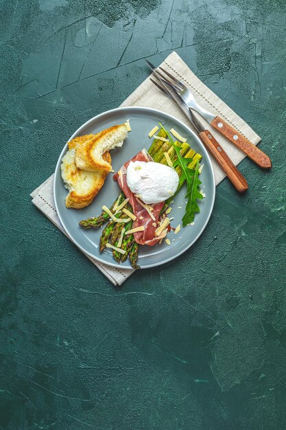 Des asperges vertes et des œufs pochés bouillis avec du jambon et du parmesan parmigiano haché sur une assiette vert foncé sur fond de béton