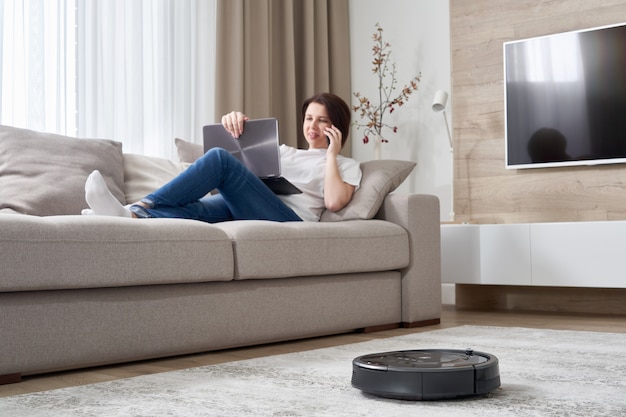 Aspirateur robot nettoyant la chambre pendant que la femme se repose sur le canapé