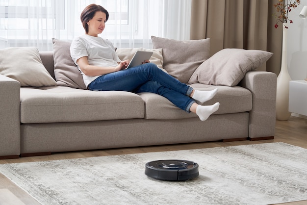 Aspirateur robot nettoyant la chambre pendant que la femme se repose sur le canapé