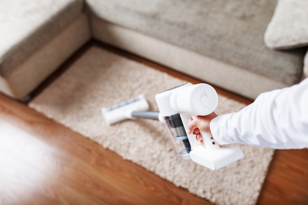 Photo aspirateur sans fil puissant avec technologie de dépoussiérage cyclonique blanc en main, nettoie le tapis dans la maison près du canapé.