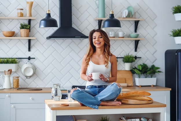 Assez heureuse femme assise sur la table dans la cuisine confortable le matin à la maison et boit du café. Détente matinale à la maison.