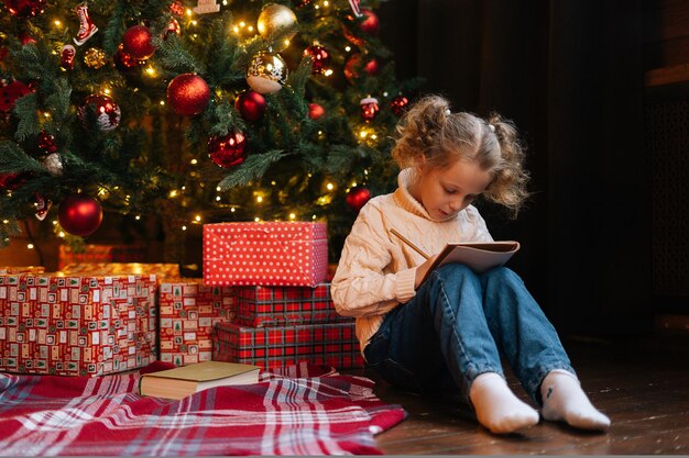 Assez heureuse petite fille blonde enfant bouclée écrivant une lettre au père noël assis sur le sol par