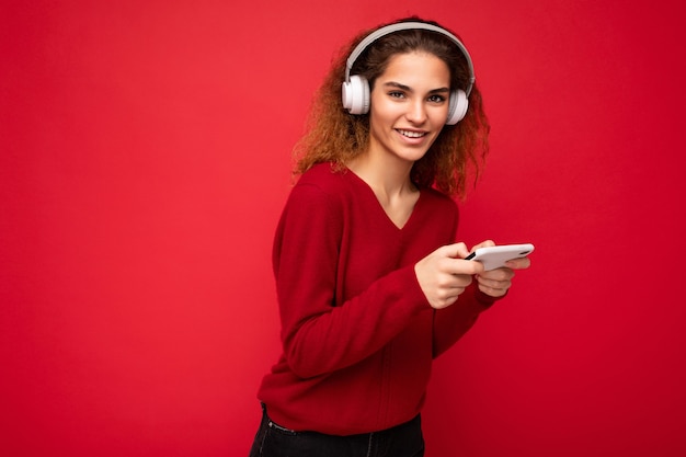 Assez heureuse souriante jeune femme brune bouclée portant un pull rouge foncé isolé sur fond rouge portant des écouteurs blancs écoutant de la musique et utilisant un smartphone en regardant la caméra.