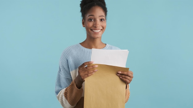 Photo assez joyeuse fille afro-américaine souriante tenant une enveloppe avec les résultats des examens regardant joyeusement à huis clos sur fond coloré