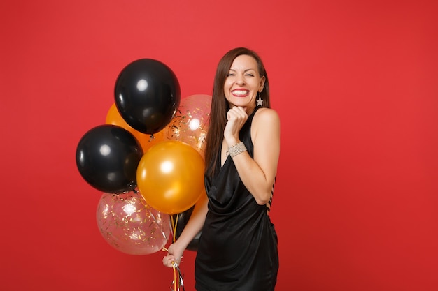 Assez joyeuse jeune fille heureuse en petite robe noire célébrant la tenue de ballons à air isolés sur fond rouge vif. Concept de fête de vacances de maquette d'anniversaire de bonne année de la journée internationale de la femme.