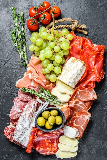 Assiette antipasti viande froide avec raisins, prosciutto, tranches de jambon, boeuf séché, salami chorizo, fuet, camembert et fromage de chèvre. Surface noire. Vue de dessus