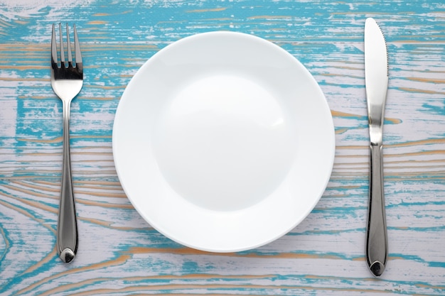 Assiette blanche vide avec une fourchette et un couteau en argent sur une table en bois bleue. Réglage du lieu du dîner. Vue de dessus.