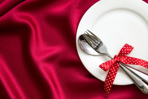 Assiette blanche vide avec une fourchette et une cuillère sur un tissu en soie rouge pour le dîner