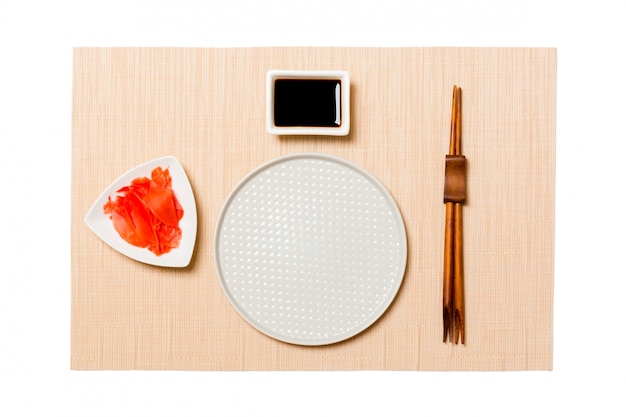 Assiette blanche vide et ronde avec des baguettes pour sushi, gingembre et sauce soja sur tapis de sushi brun. Vue de dessus avec espace de copie