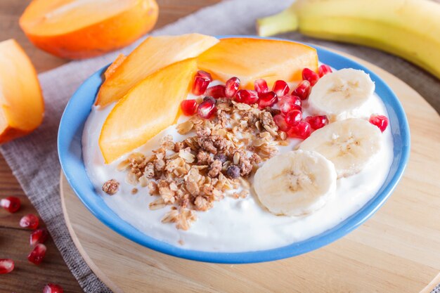 Une assiette bleue avec du yaourt grec, granola, kaki, banane, grenade sur bois brun.