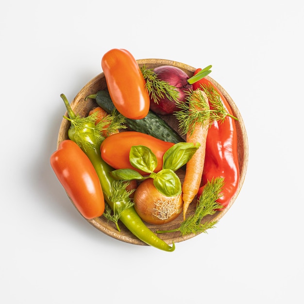 Assiette en bois avec des légumes frais poivron vert tomate oignon carotte concombre basilic aneth sur fond blanc vue de dessus concept rustique