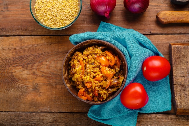 Une assiette de boulgour avec des légumes et des champignons à côté d'oignons et de tomates et une tasse de boulgour sur une table en bois