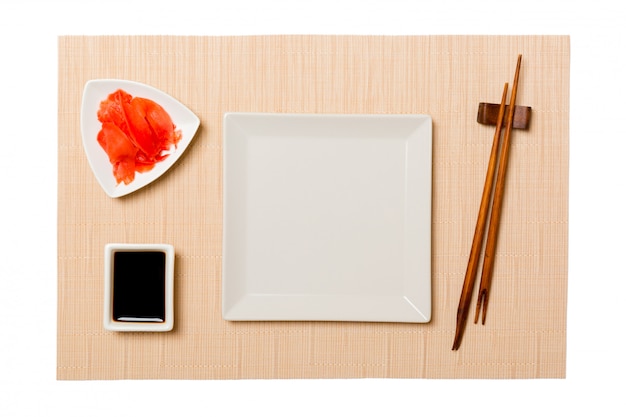 Assiette carrée blanche vide avec des baguettes pour sushi et sauce soja, gingembre sur fond de tapis de sushi marron.