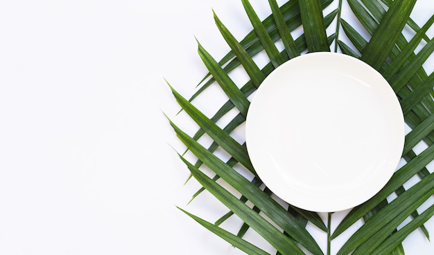 Assiette en céramique blanche vide sur des feuilles de palmier tropical sur fond blanc. Vue de dessus