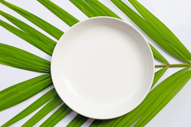 Assiette en céramique blanche vide sur les feuilles de palmier tropical sur fond blanc.