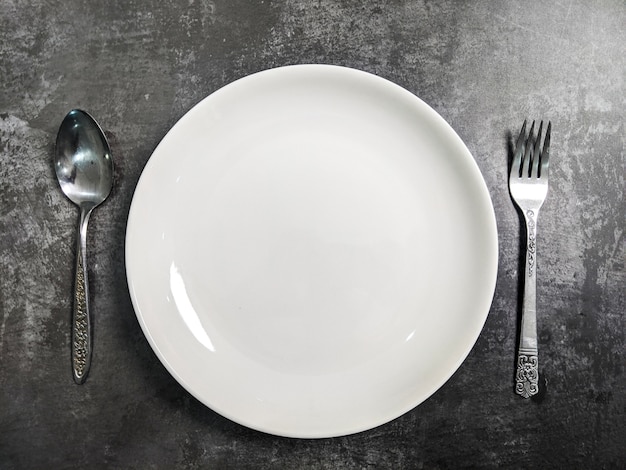 Assiette en céramique blanche vide avec fourchette et cuillère sur fond de table en béton de pierre grise. Copiez l'espace. Concept de recette de menu