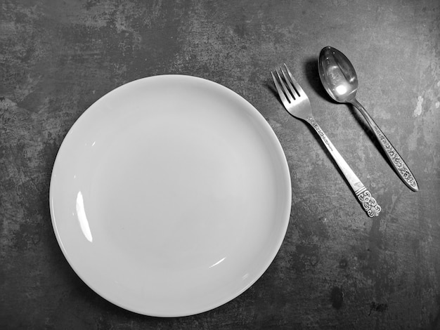 Assiette en céramique blanche vide avec fourchette et cuillère sur fond de table en béton de pierre grise. Copiez l'espace. Concept de recette de menu