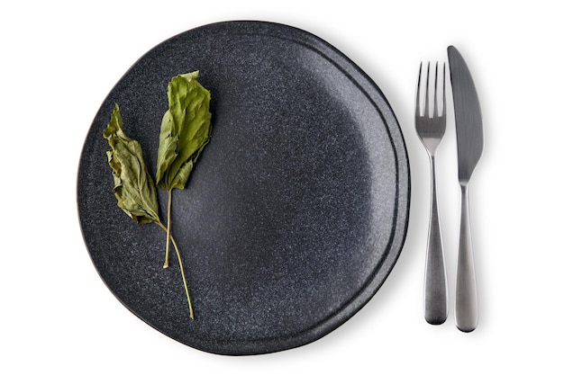 Assiette en céramique noire vide décorée de feuilles de cassis séchées avec des couverts isolés sur fond blanc. Disposition des menus