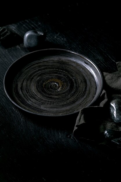 Assiette en céramique noire vide avec des pierres noires autour sur une serviette textile sur fond de bois noir.