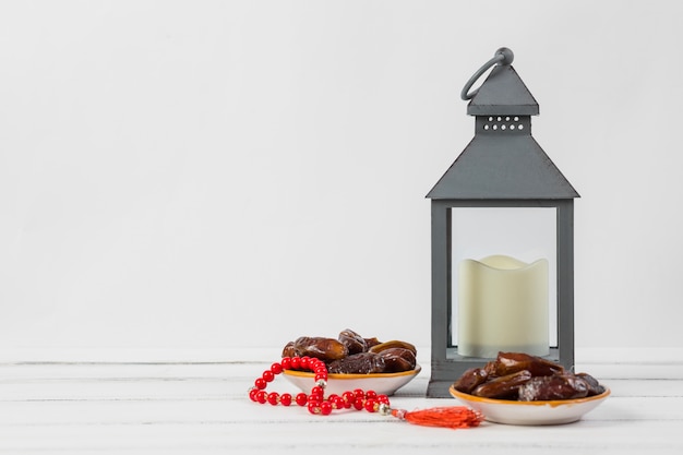 Assiette de dattes juteuses avec des perles de prière rouges et une bougie dans un support de lanterne sur fond blanc