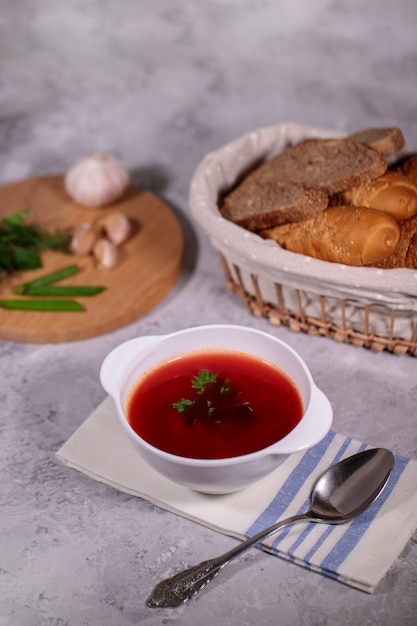 Une assiette avec du bortsch à la betterave rouge sur la table, à côté du plateau, contient du persil, de l'aneth, des oignons verts et de l'ail et un panier avec du pain. Déjeuner savoureux et copieux.