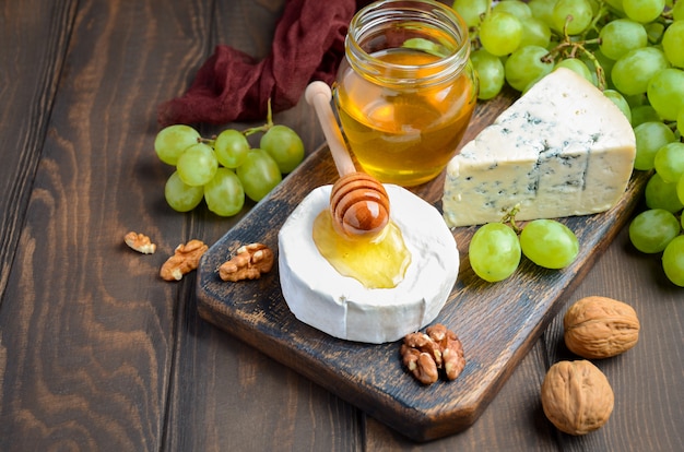 Assiette de fromages aux raisins, miel et noix sur fond sombre.